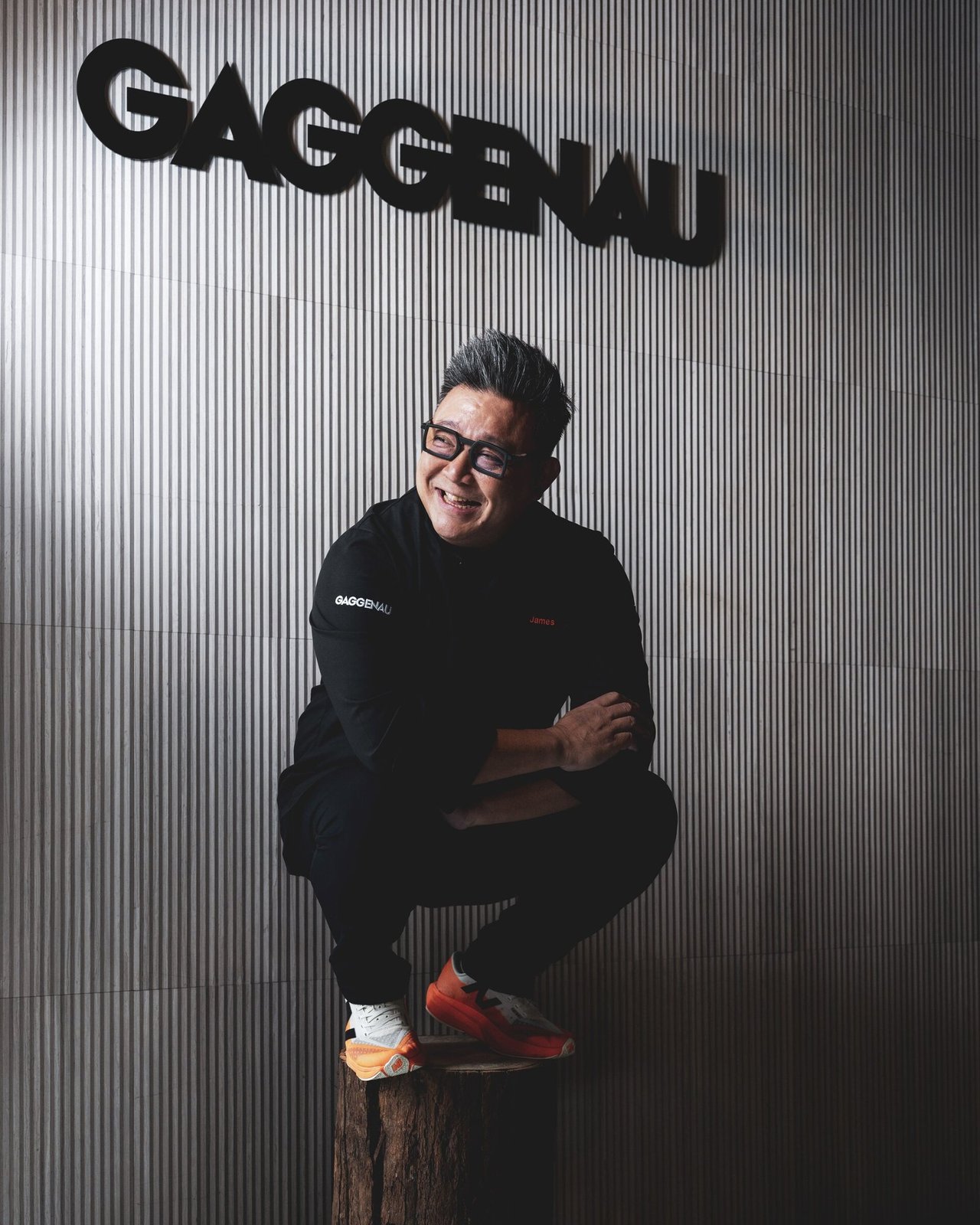 James Won seated on the wooden stump in front of the GAGGENAU sign — tighter frame focusing on the figure and brand mark.
