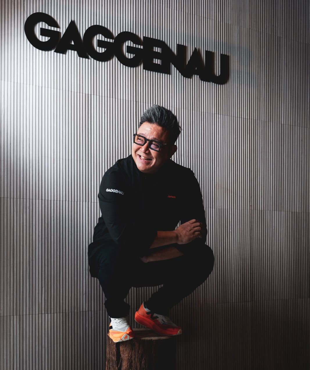 James Won on a wooden stump in front of the GAGGENAU sign — wider frame showing the full Inspiration Haus wall, orange Asics sneakers visible.