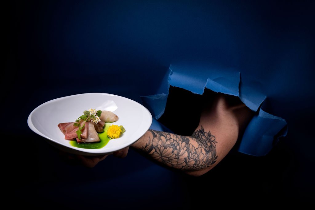 A composed course on a white plate held by a tattooed arm through torn dark-blue paper.