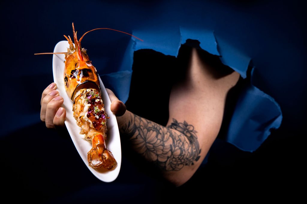 A whole glazed langoustine on a white oval plate, held by a tattooed arm emerging through torn dark-blue paper.