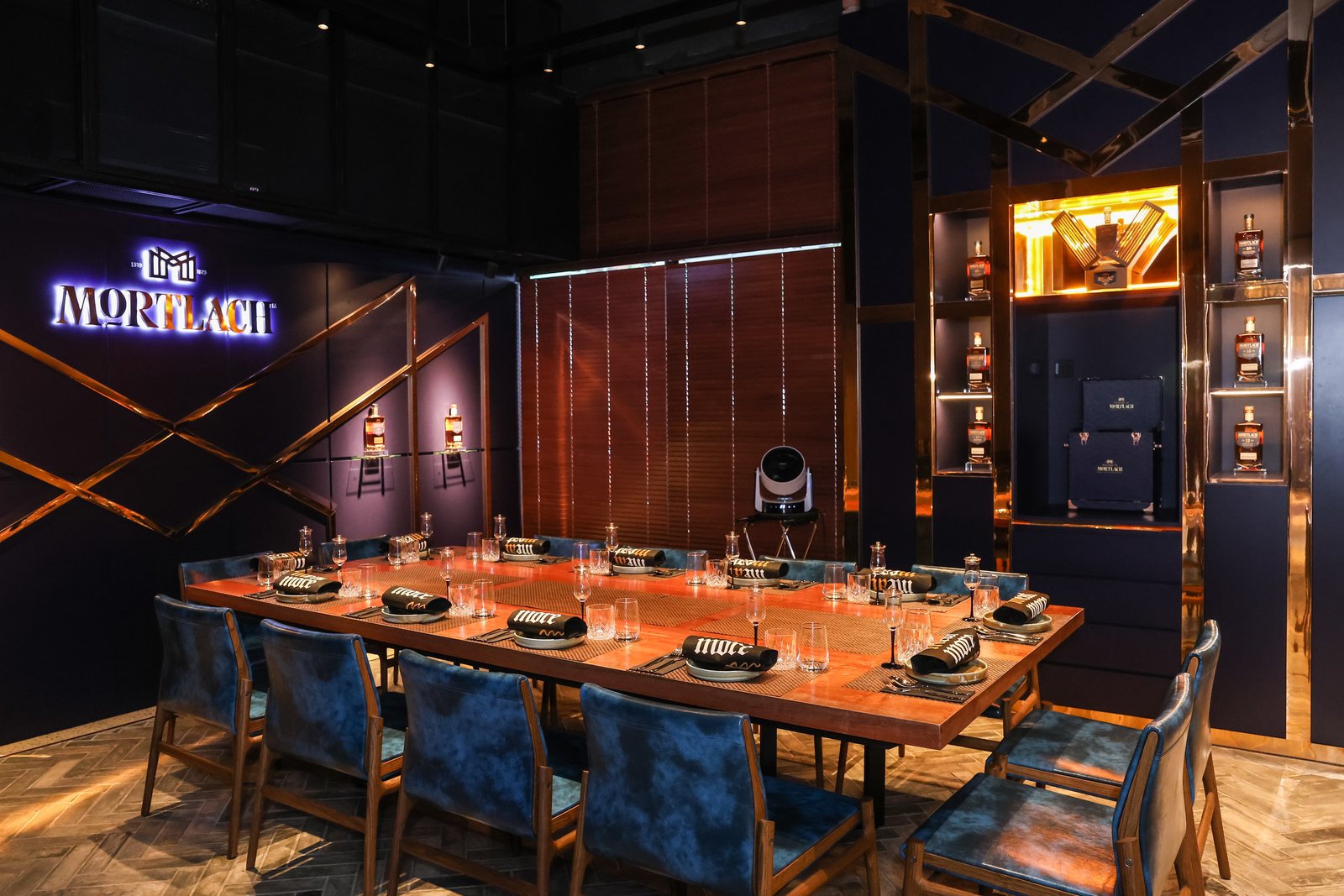 The Mortlach Room at MeatMore — a long communal wooden table set for twelve beneath a blue neon MORTLACH sign, with dark panelled walls and a bottle display.