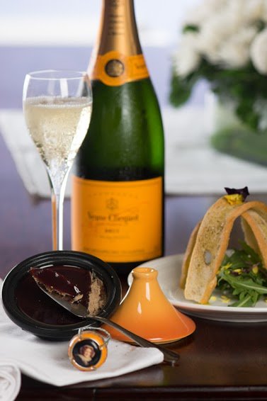 Veuve Clicquot Brut bottle with champagne flute, alongside a small chocolate dome dessert and an arancini-like savoury course in cone form, white roses behind.