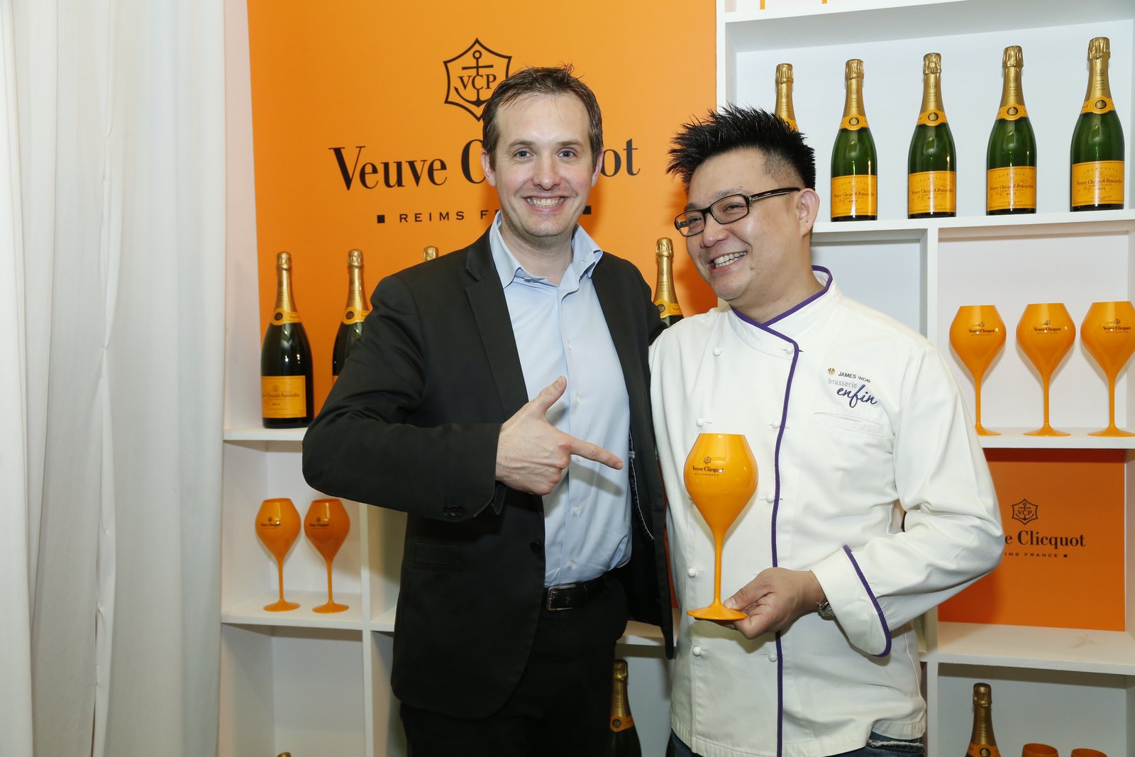 James Won and the Veuve Clicquot Maison representative at Brasserie Enfin, late May 2015 — the orange Veuve Clicquot Reims France logo wall behind, bottles and orange flutes shelved alongside.