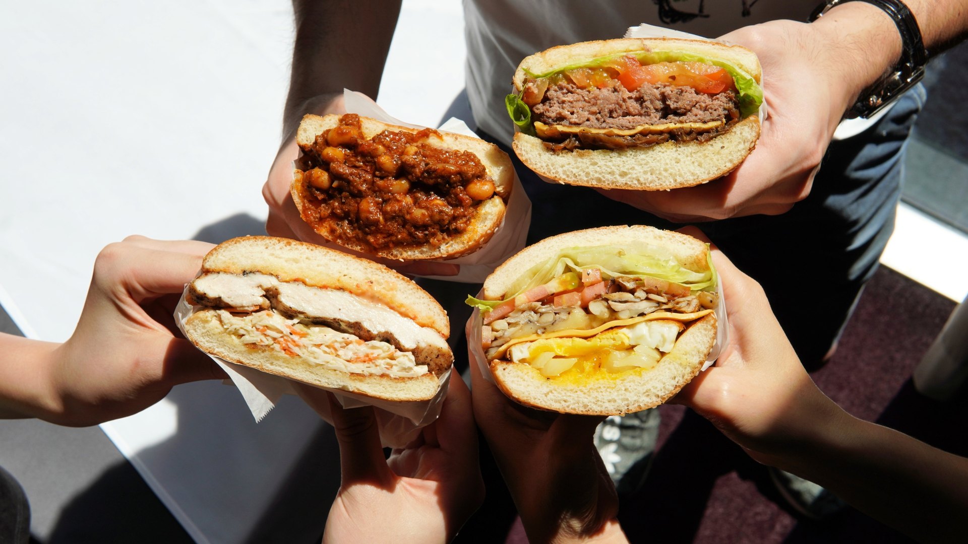 Four hands holding four different burgers and sandwiches against bright daylight — chilli con carne, beef burger, fried chicken, and a vegetarian sandwich.