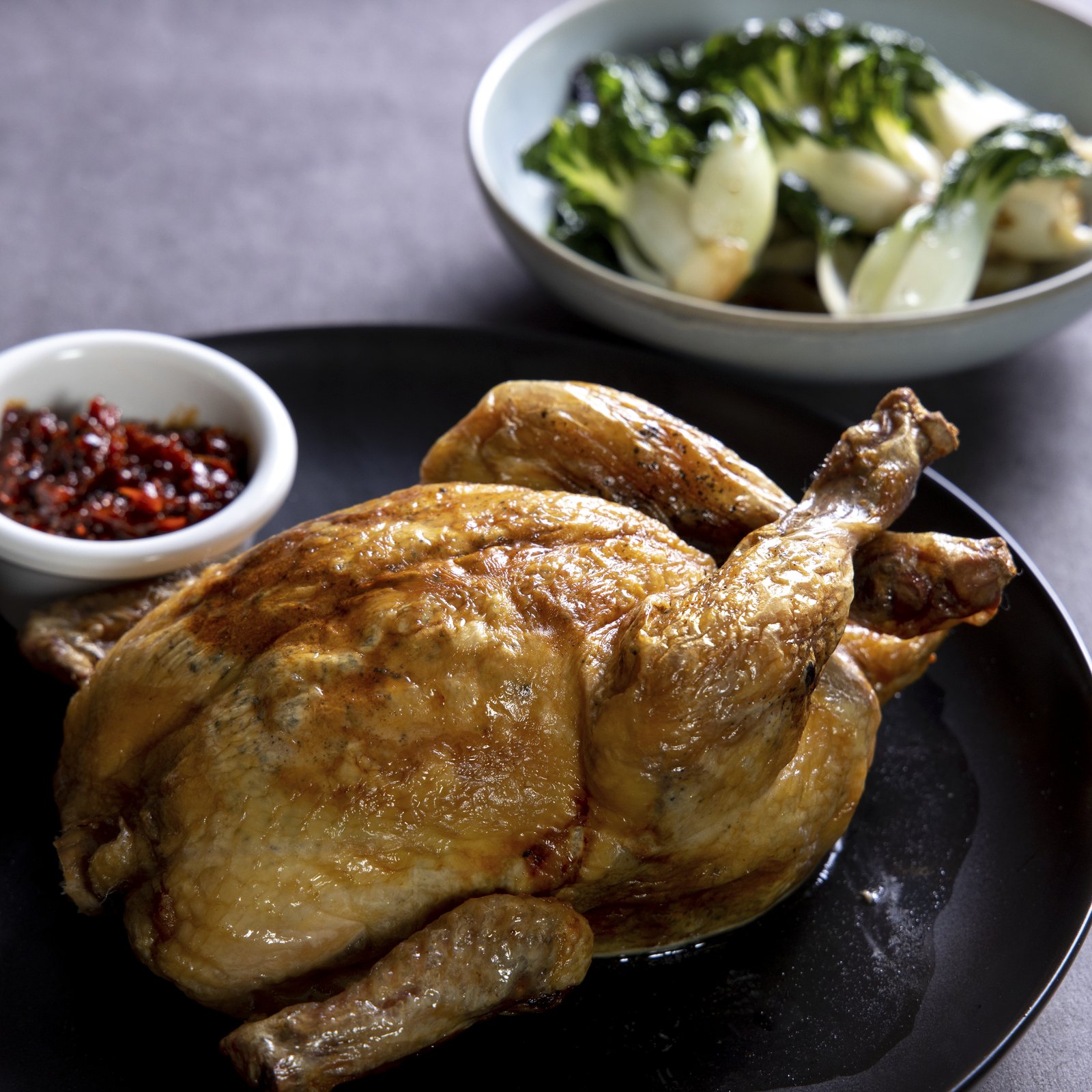 Bouchon Enfin whole roast chicken — golden-brown, served whole with bok choy and a small bowl of chilli condiment.